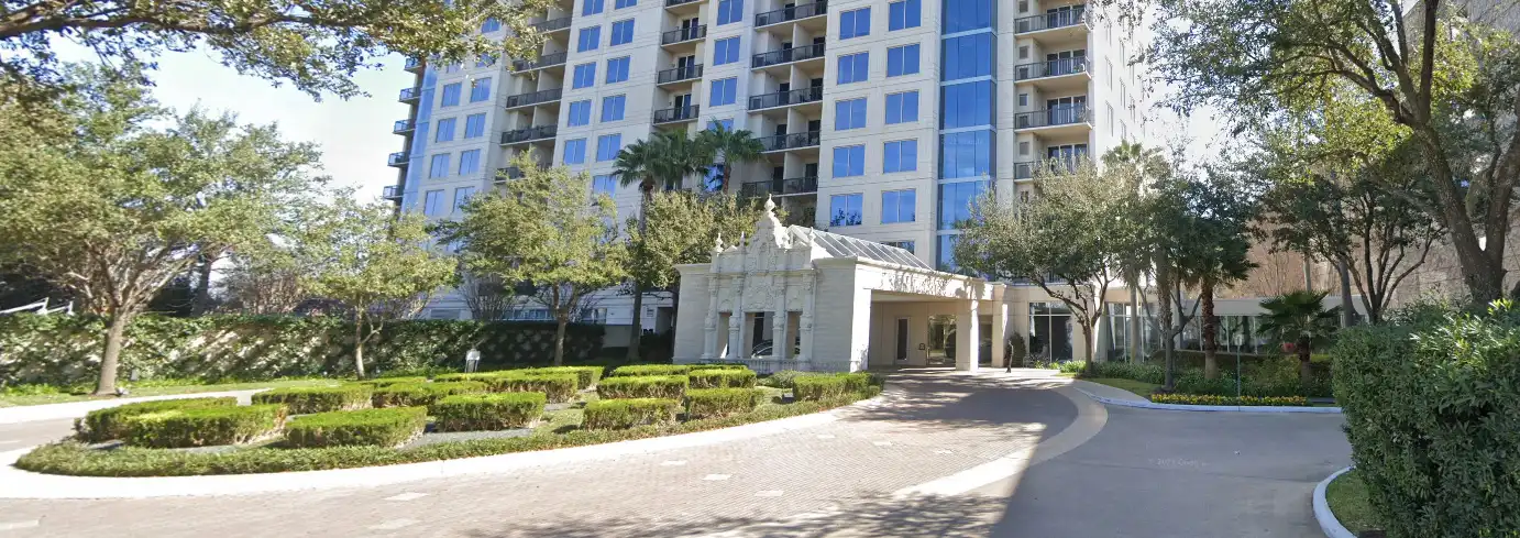 A tall, modern apartment building with balconies stands behind a circular driveway lined with manicured bushes, trees, and neatly trimmed hedges under a partly cloudy sky.