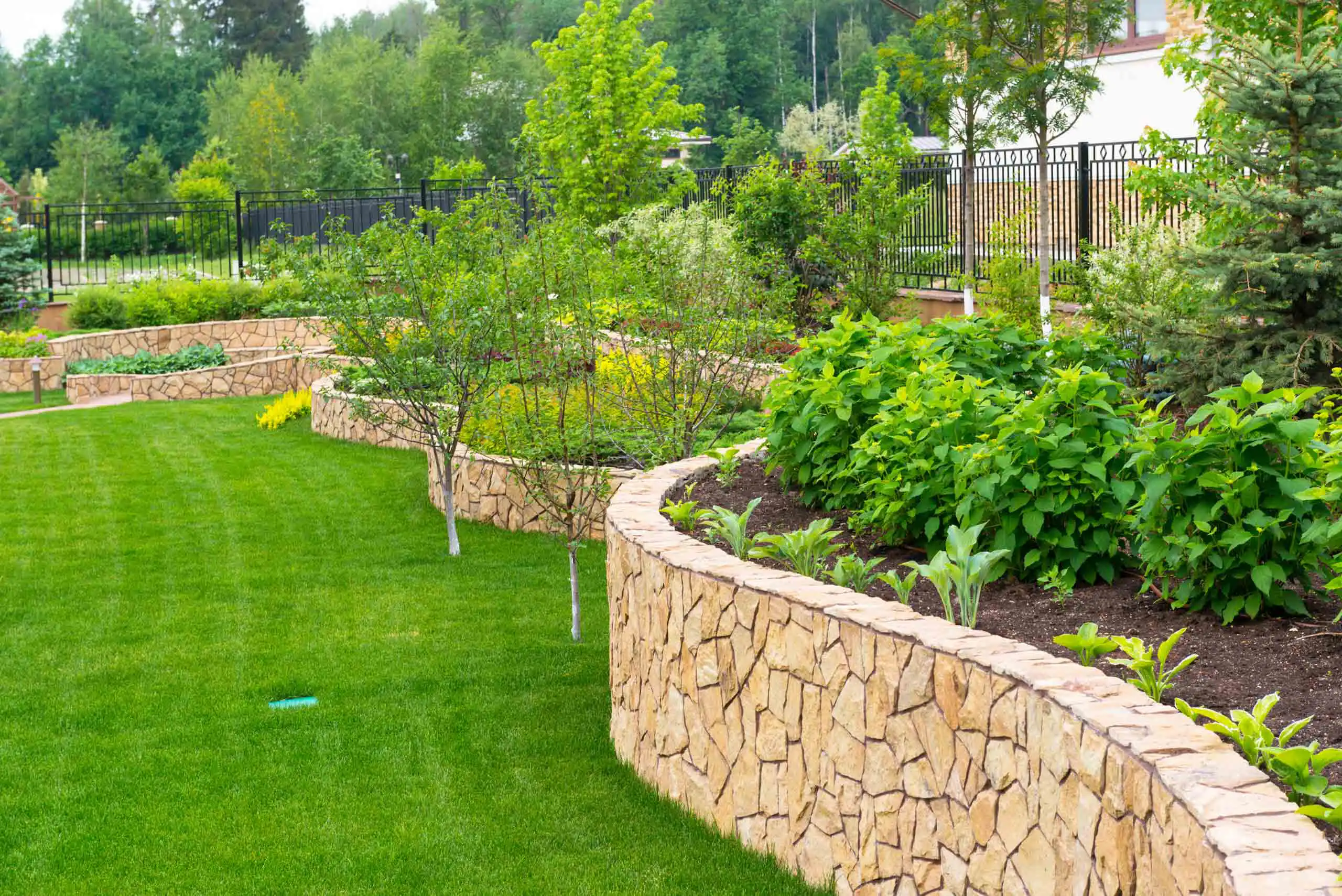 A landscaped garden with neatly trimmed grass and raised stone flowerbeds filled with green plants and small trees, surrounded by a black metal fence and lush greenery in the background.